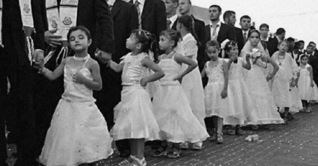 A line of young girls in white dresses standing beside adult men in black suits during a traditional ceremony in Iraq.