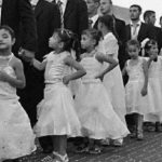 A line of young girls in white dresses standing beside adult men in black suits during a traditional ceremony in Iraq.