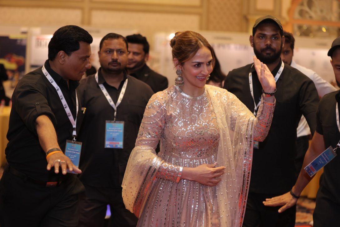 A woman in a sparkly traditional outfit waves while being escorted by several security personnel at an event.