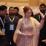 A woman in a sparkly traditional outfit waves while being escorted by several security personnel at an event.