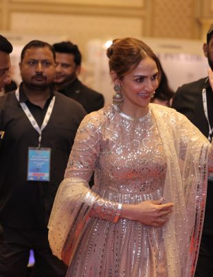 A woman in a sparkly traditional outfit waves while being escorted by several security personnel at an event.